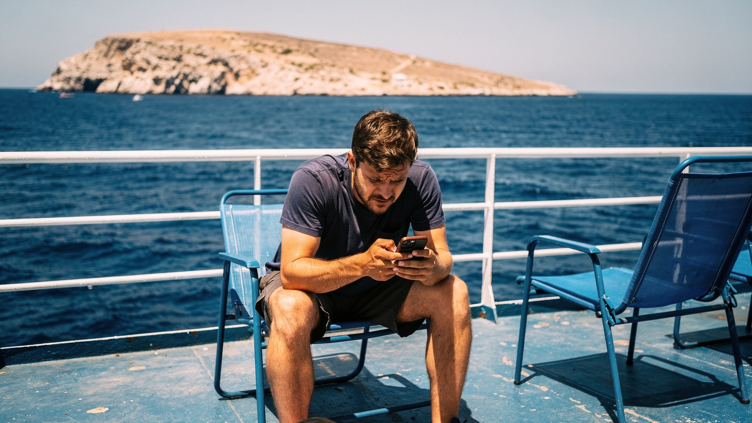 A frustrated traveler sitting on an outdoor ferry deck staring at a smartphone screen.