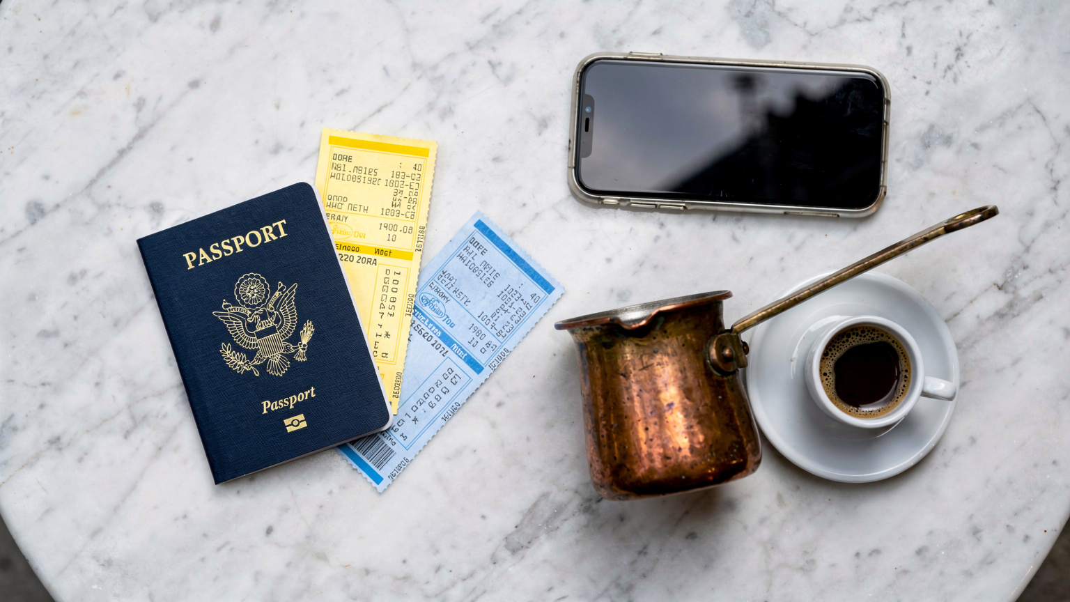 An overhead flat lay of a US passport, printed ferry tickets, a smartphone, and a cup of Greek coffee on a cafe table.