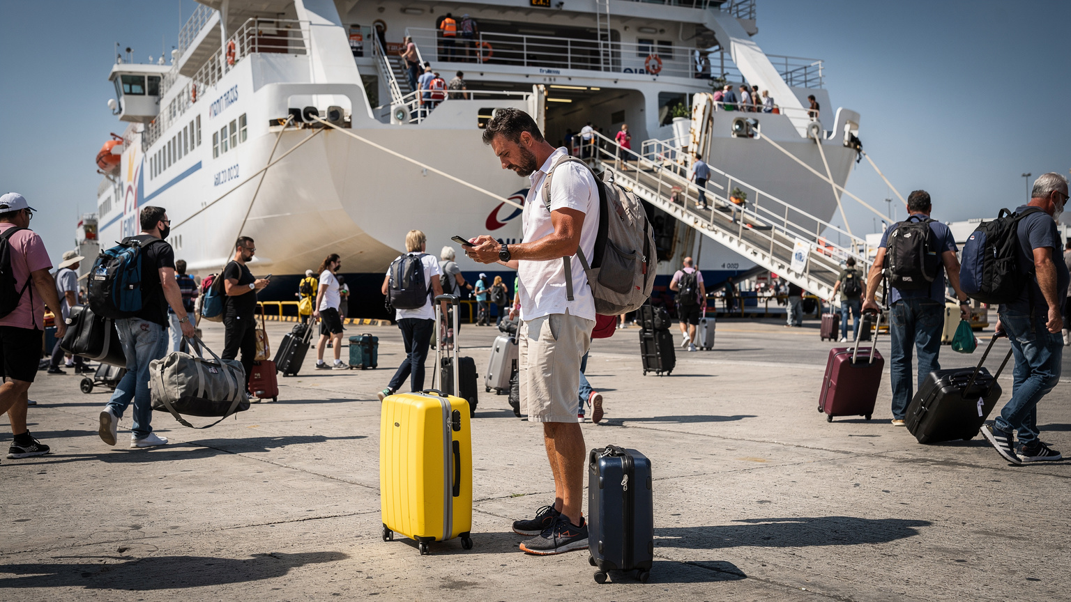 A digital nomad waiting at Piraeus port gate E7 with luggage and a smartphone in hand.