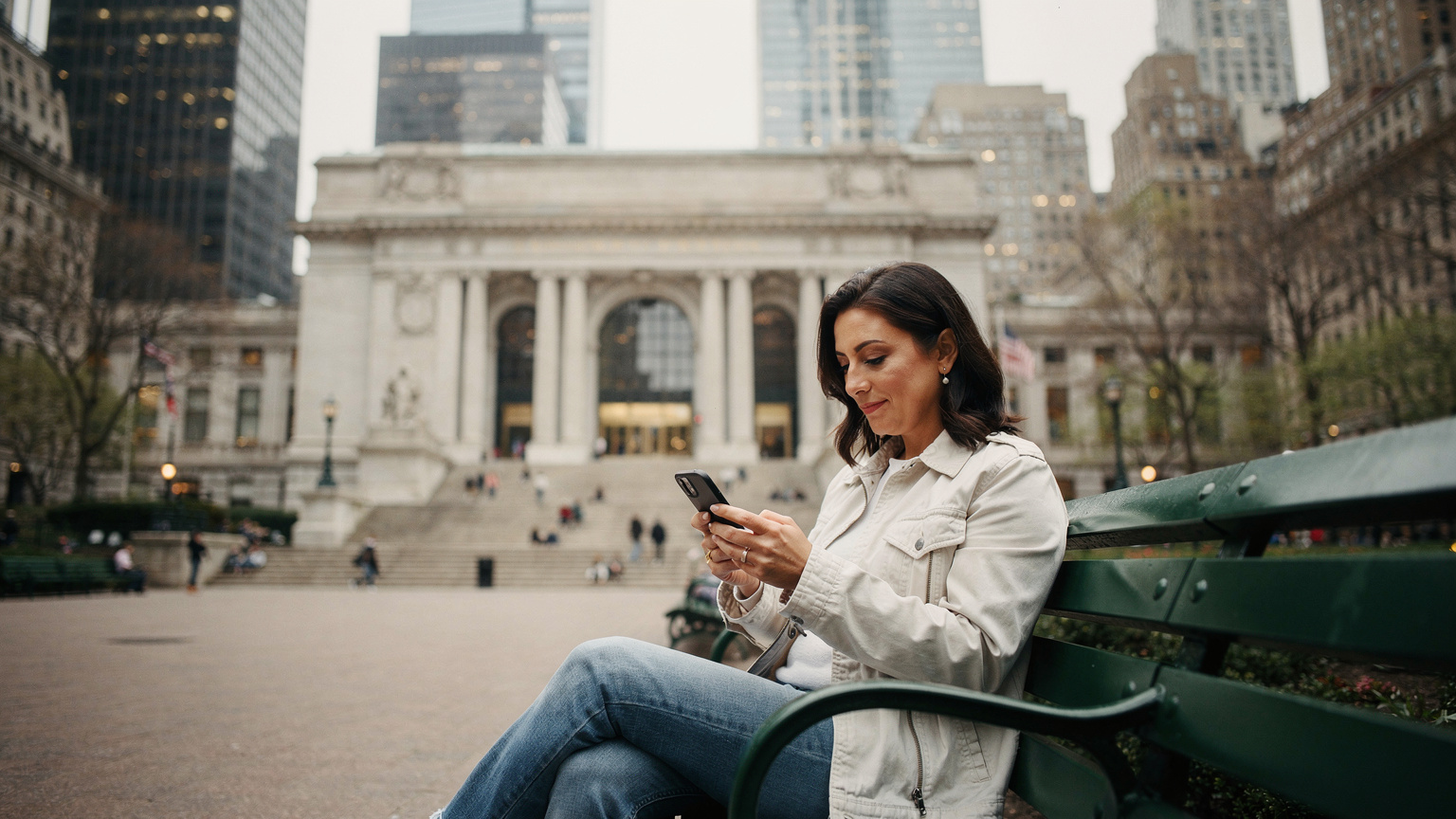 Μια γυναίκα κάθεται σε ένα παγκάκι στο Bryant Park της Νέας Υόρκης, κρατώντας ένα τηλέφωνο, με το γραφικό σκηνικό της βιβλιοθήκης και των ουρανοξυστών στο βάθος.