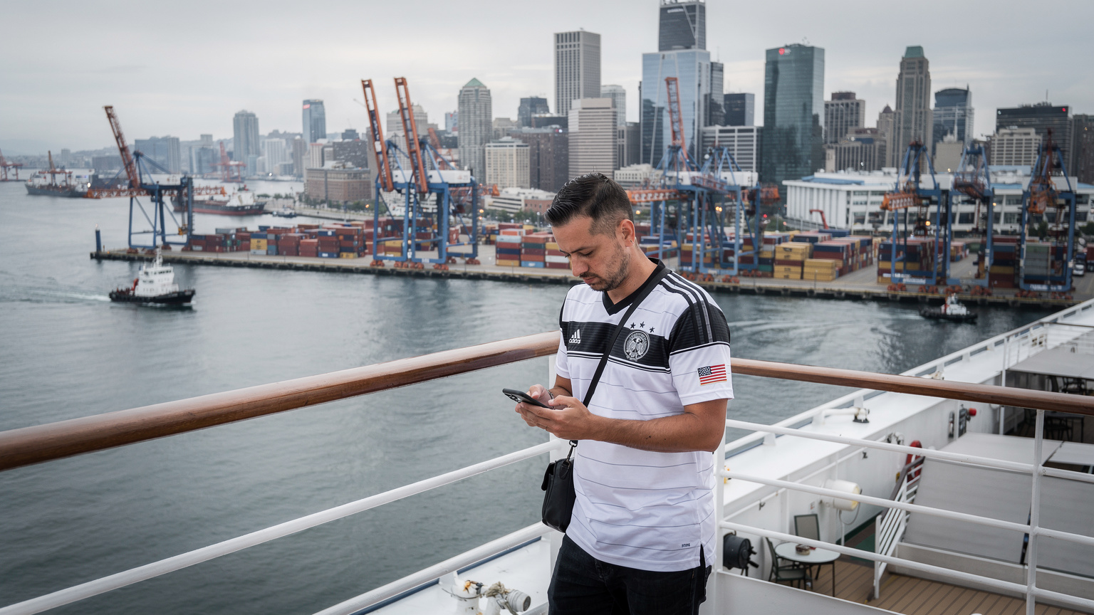Ein Reisender, der ein Deutschland-Fußballtrikot trägt, steht auf dem Oberdeck eines großen Kreuzfahrtschiffs und blickt auf die Küstenlinie einer belebten nordamerikanischen Stadt. Er hält sein Smartphone in der Hand und schaut darauf, während im Hintergrund ein Hafen mit Containern und Kränen zu sehen ist. Der Himmel ist leicht bewölkt, die Szene wirkt authentisch und ungestellt.