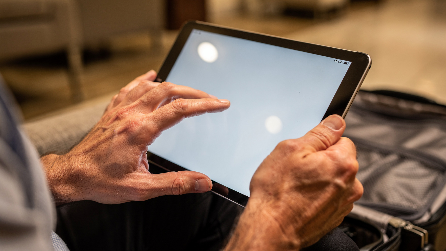 Nahaufnahme der Hände eines Vaters, der auf einem Tablet für seine Tochter offline verfügbare Filme und Serien über eine Streaming-App herunterlädt. Im Hintergrund sind angedeutet Reisegepäck und eine Hotellobby zu sehen, die Lichtstimmung ist neutral und funktional.