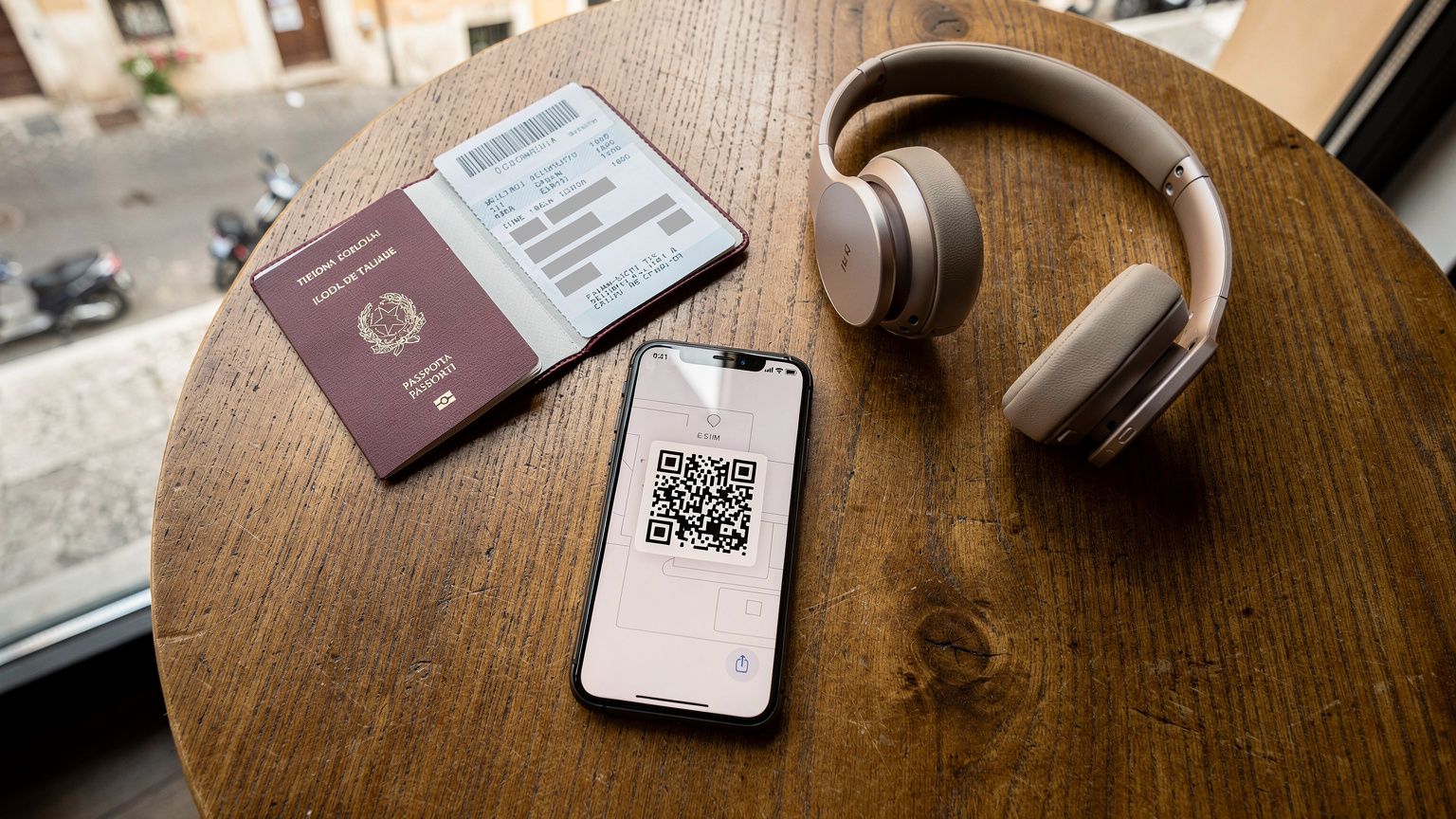 Una prospettiva dall'alto di un tavolino da caffè in una caffetteria luminosa a Roma, con un passaporto italiano, un biglietto aereo, un paio di cuffie e uno smartphone che mostra l'interfaccia di attivazione di una eSIM con un QR code visibile.
