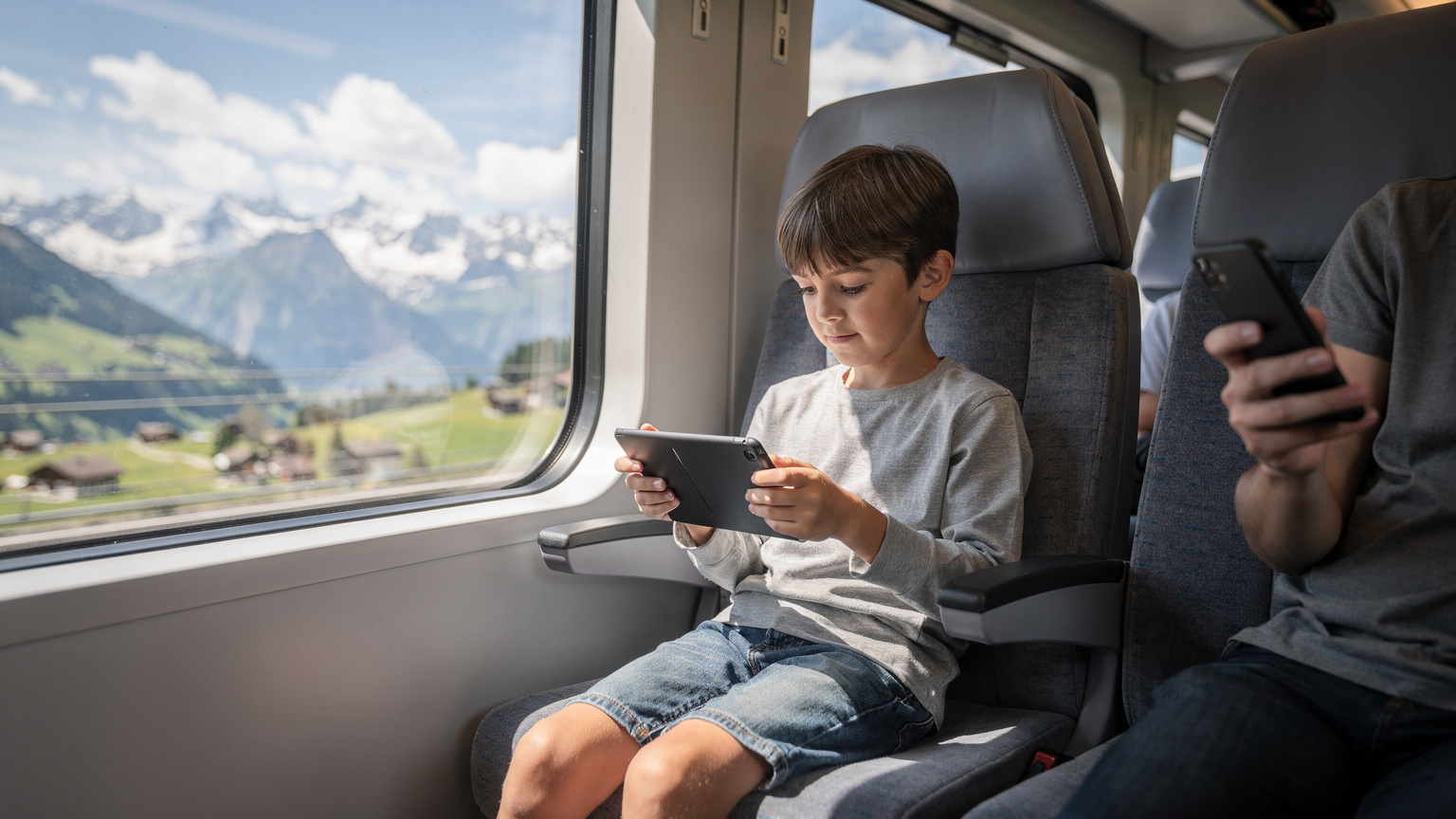 Ein Kind spielt mit einem Tablet, während ein Elternteil im Hintergrund in einem Zug durch die Schweizer Alpen auf sein Handy schaut.