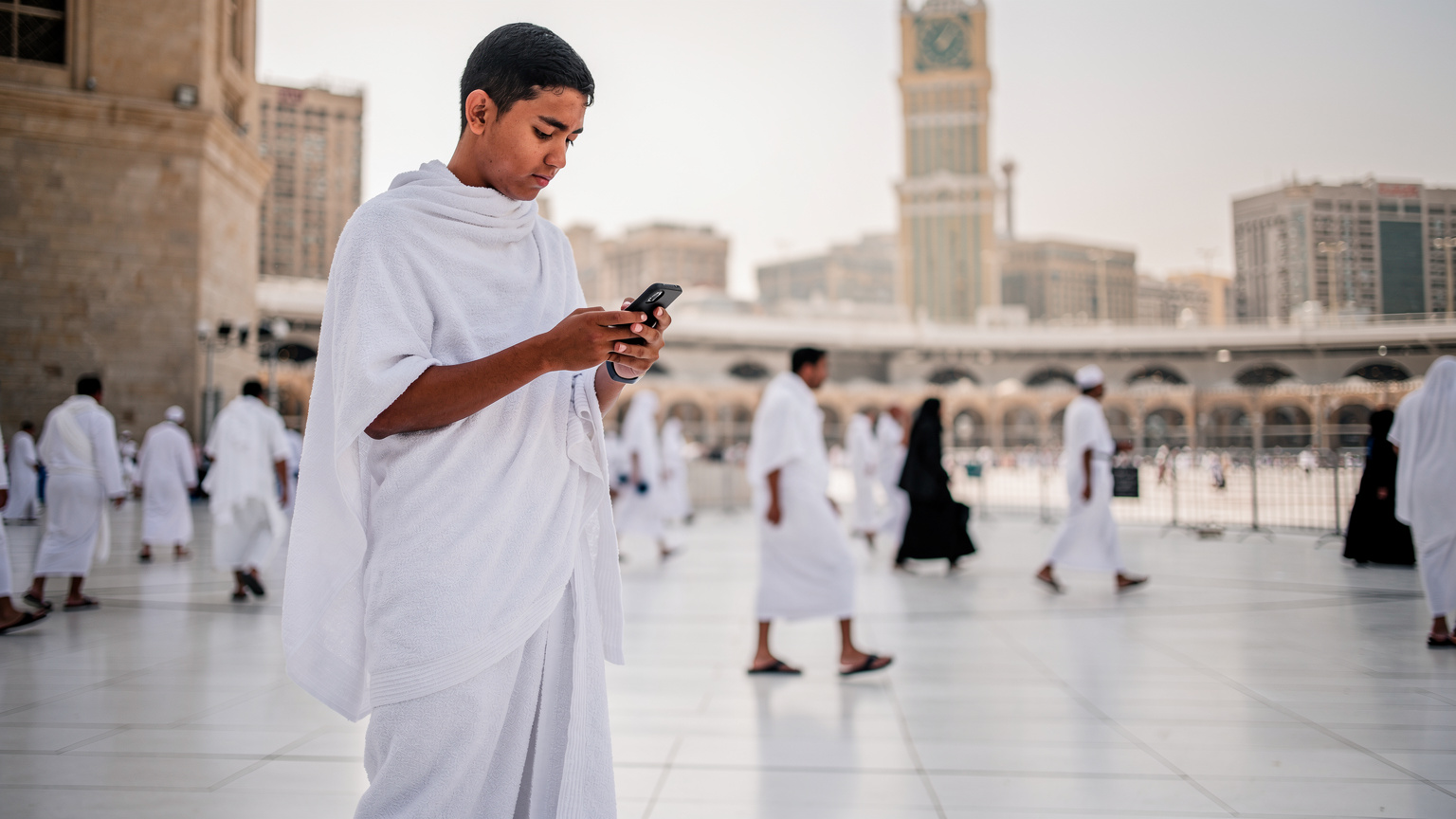 Seorang jamaah haji pria muda sedang menatap serius ponselnya, menggunakan aplikasi navigasi di tengah kerumunan di dekat Kompleks Abraj Al-Bait di Makkah.