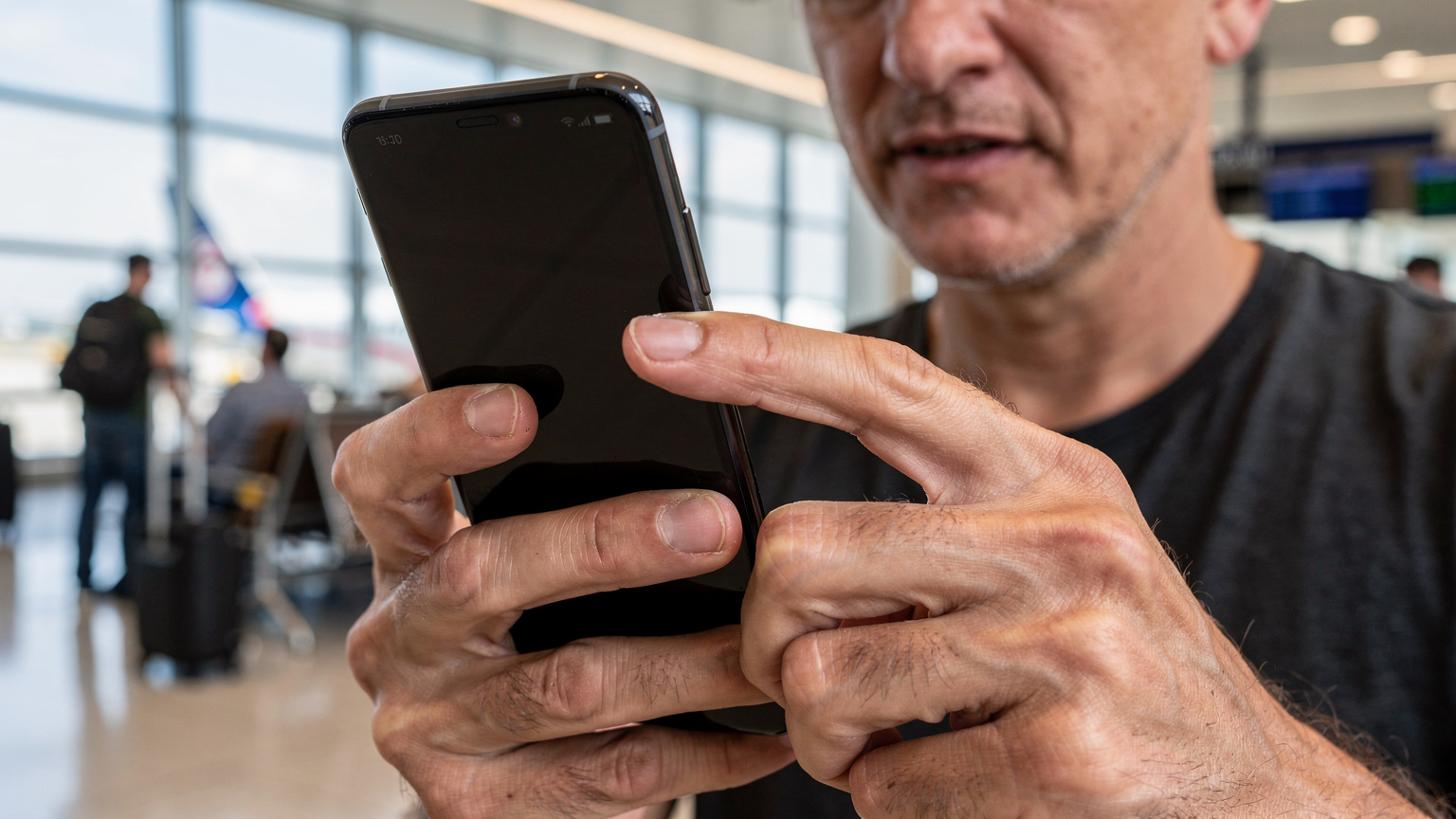 En reisende sjekker forsiktig telefonens skjerm for nettverksinnstillinger på en travel flyplassgate, med bagasje og andre passasjerer i bakgrunnen.