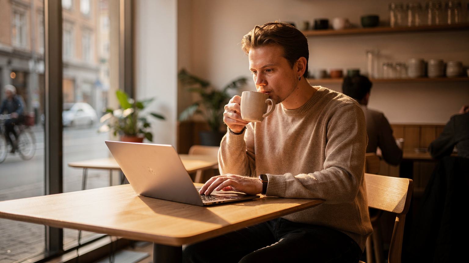 En person nipper til kaffe på en moderne, minimalistisk kafé i Oslo, med en bærbar PC åpen på bordet, som symboliserer fjernarbeid eller digitalt nomadeliv.