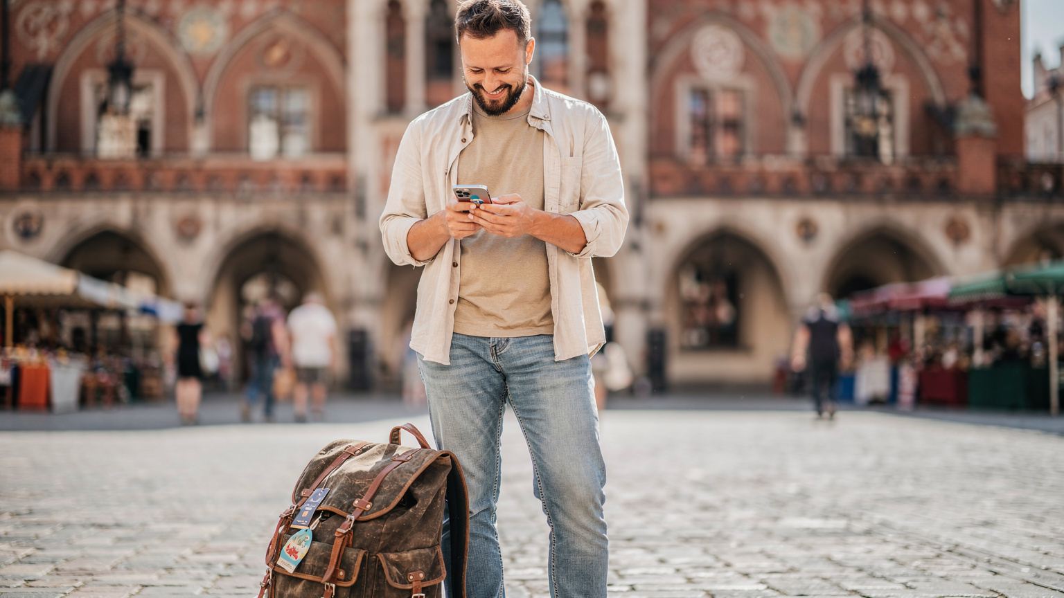 Un homme souriant et décontracté, sac à dos à ses pieds, utilise son smartphone pour recharger son forfait eSIM via une application, devant la façade colorée d'un bâtiment historique de marché à Cracovie, en Pologne, sous un soleil éclatant.