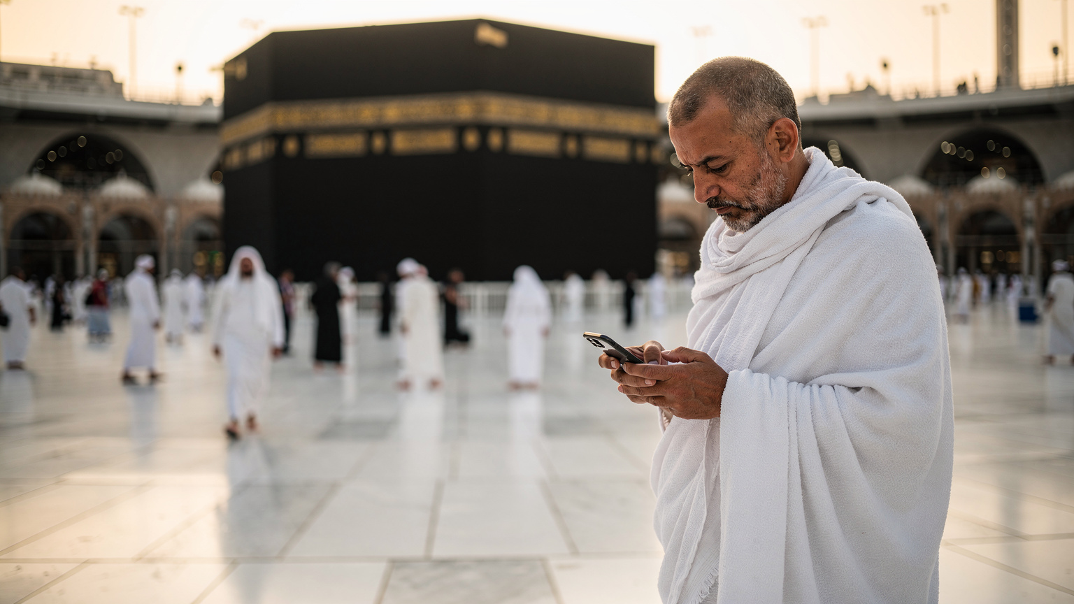 حاج يرتدي إحرامه يتفحص هاتفه الذكي بحرص أمام الكعبة المشرفة في المسجد الحرام بمكة المكرمة، وقت الفجر، بضوء ذهبي ناعم.