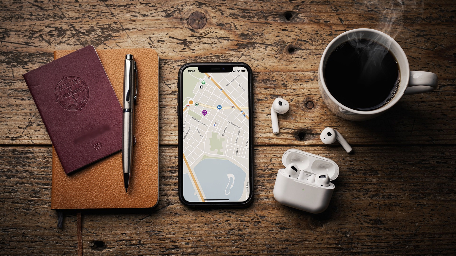 Uma vista aérea de um conjunto de itens de viagem arrumados sobre uma mesa de madeira: um smartphone com um mapa aberto, um passaporte português, um fone de ouvido sem fio, um caderno de viagem e uma caneta, com uma xícara de café ao lado.