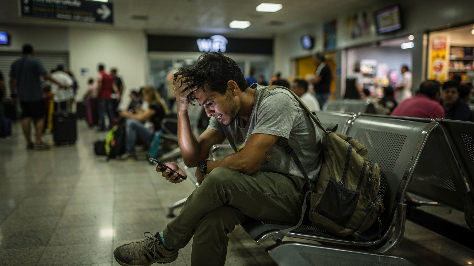 Una persona frustrada en un aeropuerto concurrido de América Latina, posiblemente el Jorge Chávez en Lima, mirando su teléfono con un letrero de "WiFi" lejano y las manos en la cabeza, expresando desesperación.