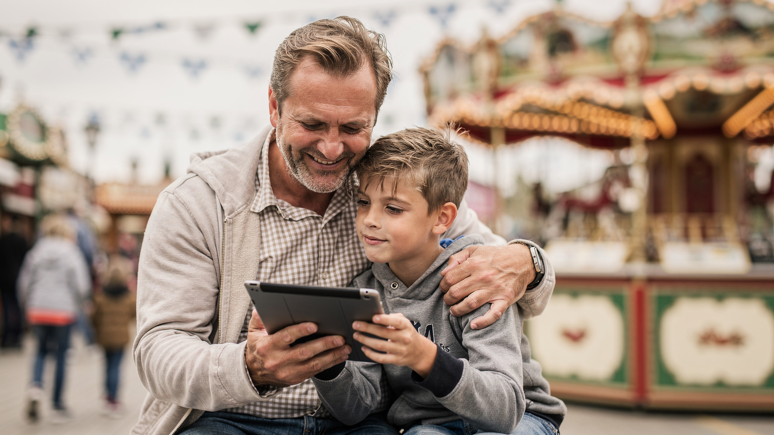 Un padre sorride mentre aiuta suo figlio a configurare un'app sul tablet, sullo sfondo la vivace atmosfera di un'area gioco all'Oktoberfest.
