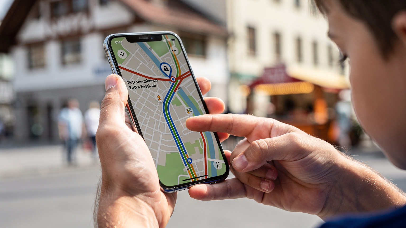 Mani di un adulto che tiene un telefono con l'app delle mappe aperta, mostrando un percorso verso l'Oktoberfest, mentre una mano di bambino indica una direzione sulla mappa.