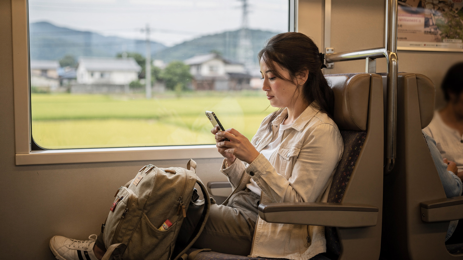 Seorang traveler sedang duduk di kursi kereta api lokal di Jepang, fokus melihat peta offline di iPhone 17, dengan latar belakang jendela yang menampilkan pemandangan pedesaan.