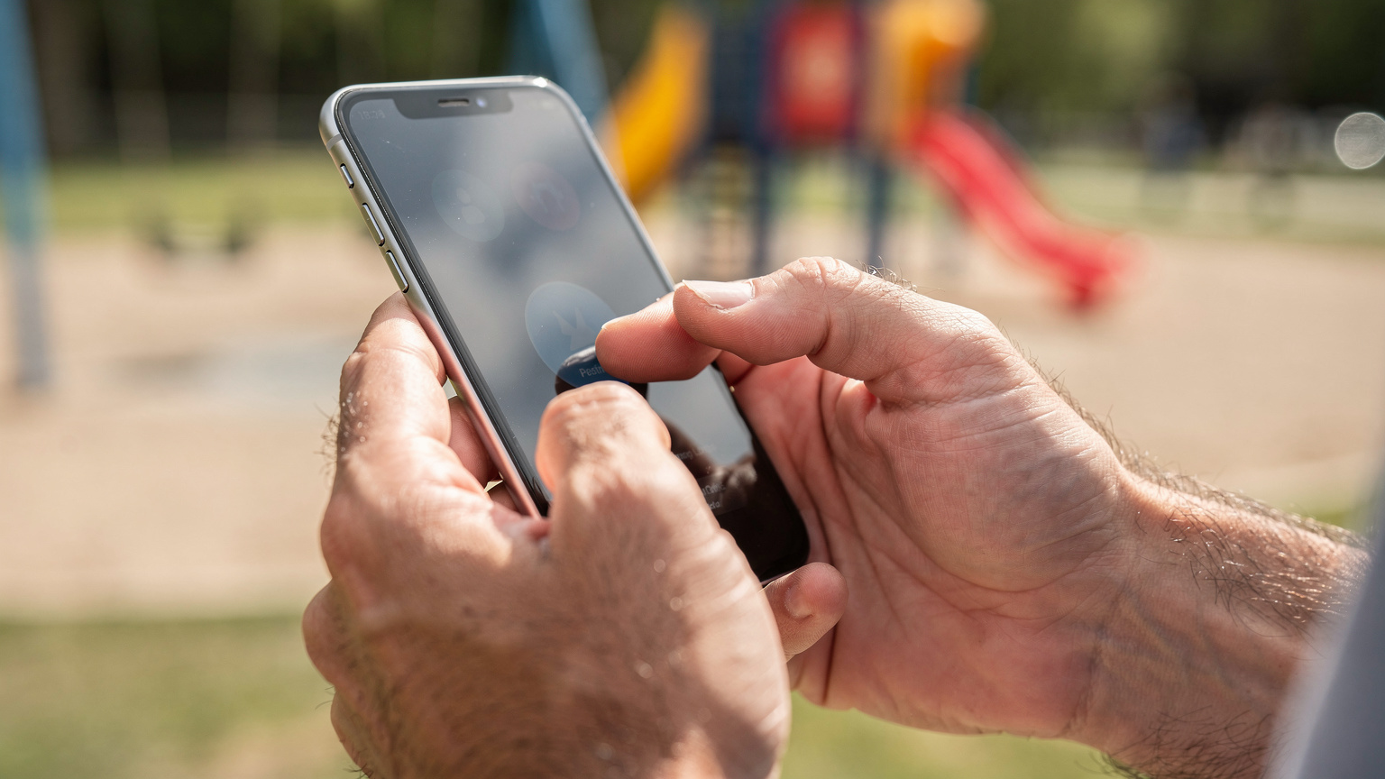 Nahaufnahme der Hände eines Vaters, der auf dem Bildschirm seines Smartphones eine Kindersicherungs-App konfiguriert, während im Hintergrund verschwommen ein Spielplatz zu sehen ist.