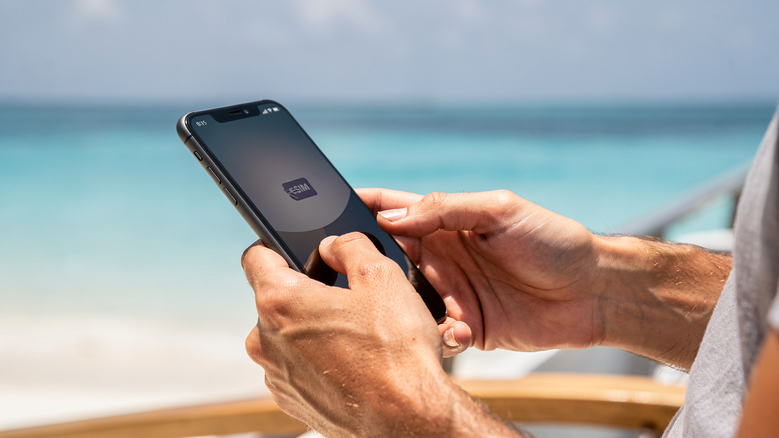 A close-up of a traveler's hands holding a smartphone, actively interacting with the screen to activate an eSIM profile, with the Maldivian ocean subtly blurred in the background.