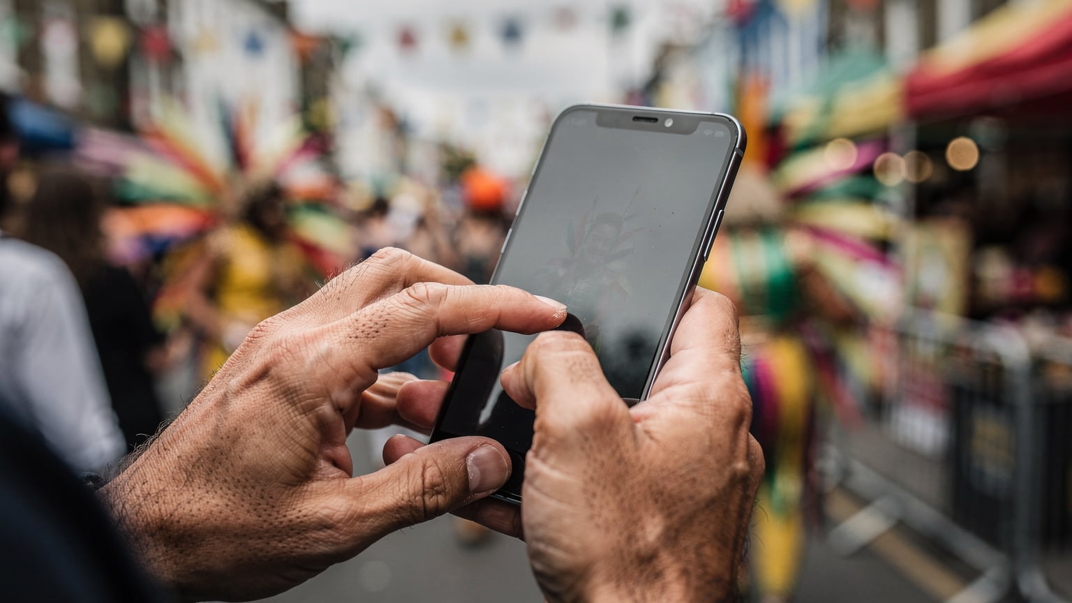 Close-up of human hands holding a smartphone with a blank or frozen screen, reflecting the frustration of a lost connection in a blurry, active street scene of Notting Hill Carnival.