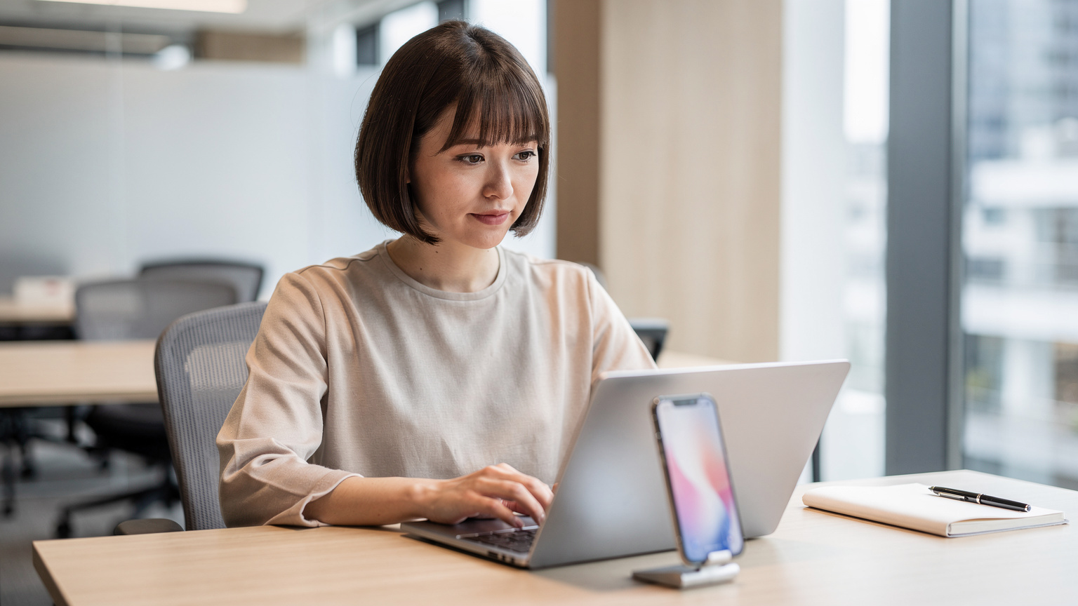 A woman sits at a modern co-working desk in the Global Business Hub Tokyo in Otemachi, her smartphone open next to a laptop, looking at the screen with focused intensity. She's wearing business casual attire.