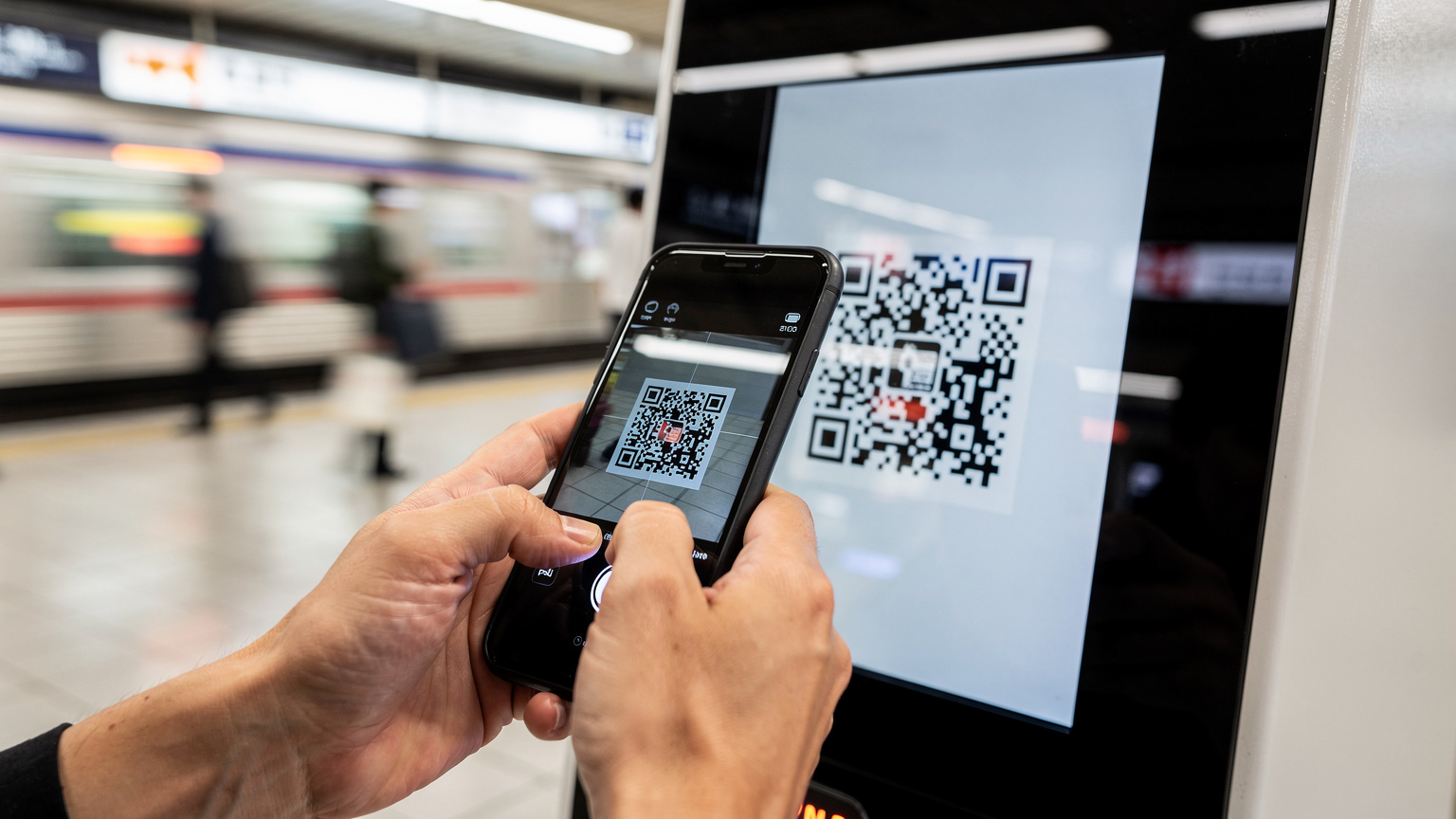 Gros plan sur des mains utilisant un smartphone pour scanner un code QR affiché sur une borne libre-service à la gare de Shinjuku, Tokyo, avec des passants flous en arrière-plan.