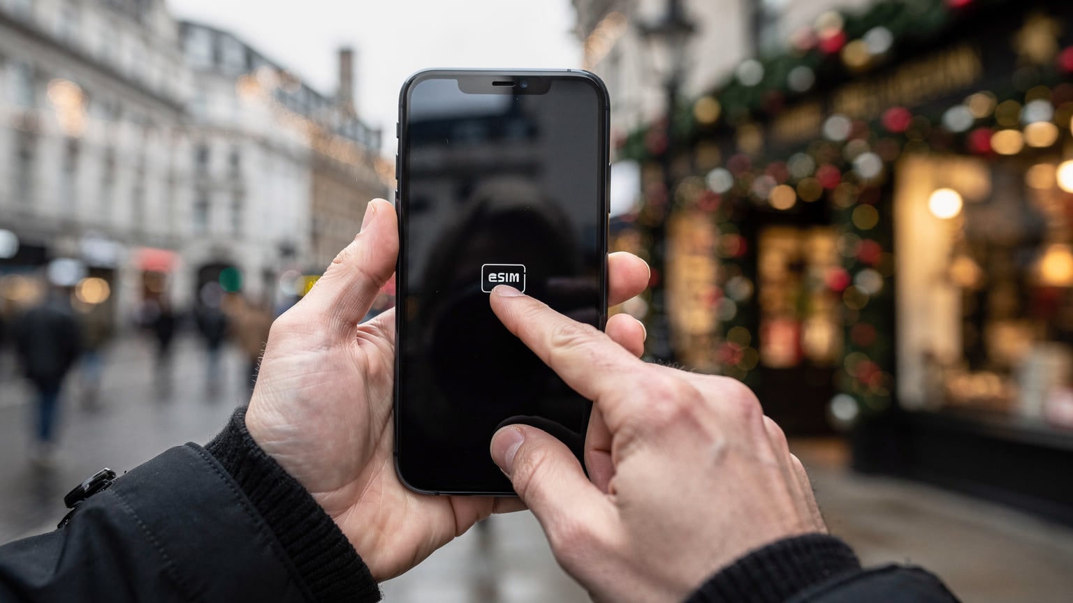 A pair of hands is shown interacting with a smartphone, setting up an eSIM profile, against the backdrop of a vibrant London street scene during the Christmas season, emphasising ease of use amidst festive distractions.