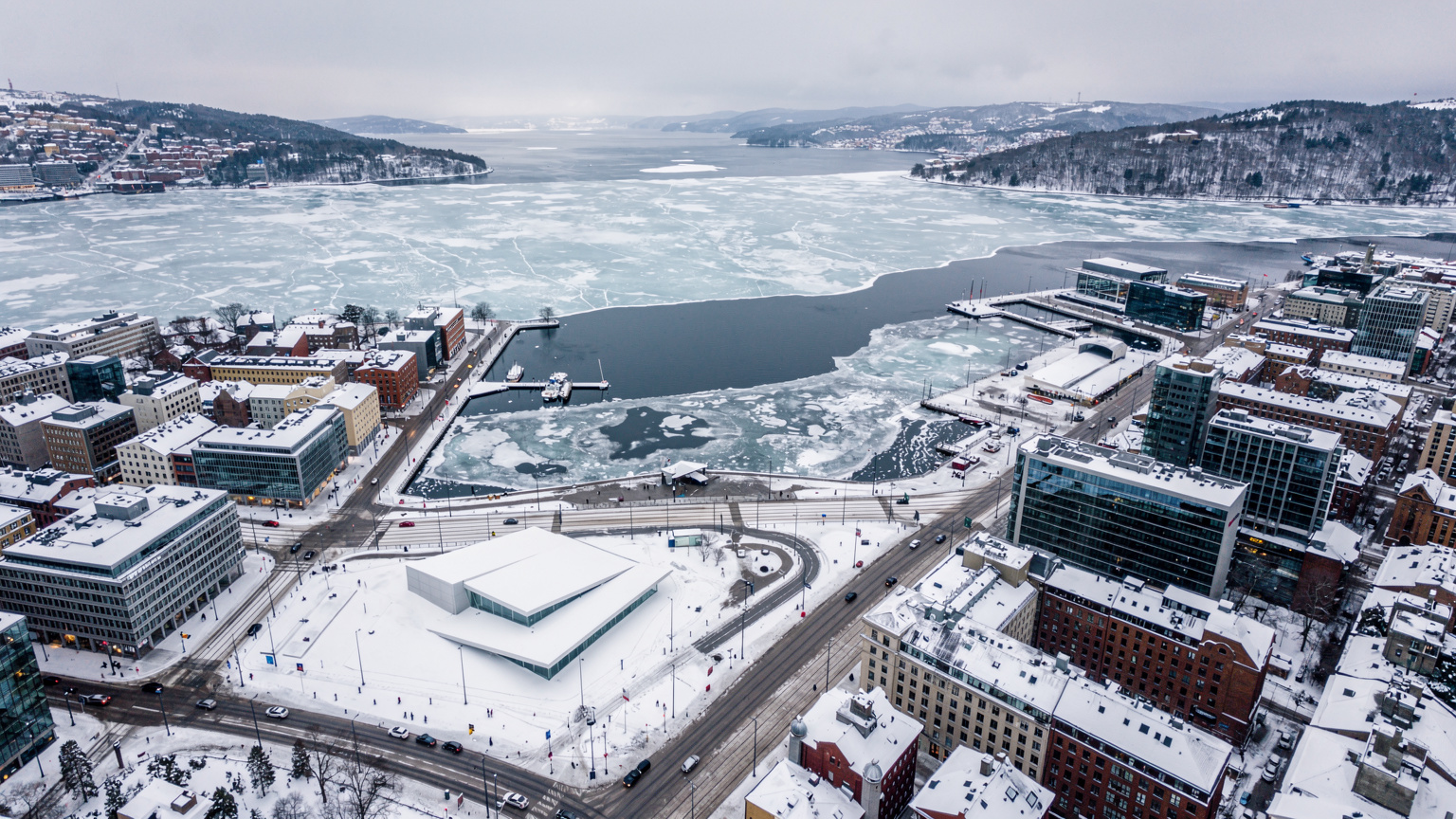 An aerial view of Oslo's city center covered in fresh snow, with the Opera House and a frozen fjord visible, under a soft, overcast winter sky.