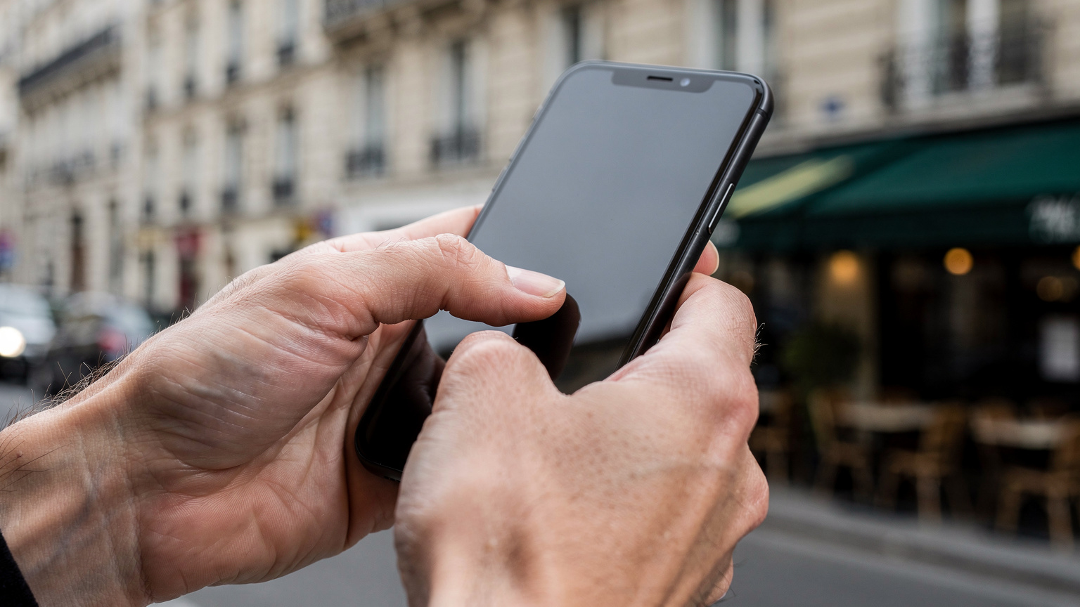 A close-up of a person's hands delicately tapping on a smartphone screen, with a blurred backdrop of a classic Parisian streetscape, suggesting active engagement with a travel app.