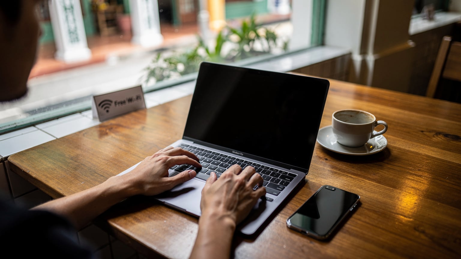 A close-up of hands typing on a laptop at a cafe in Singapore, with a small sign advertising 'Free Wi-Fi' subtly visible in the background, illustrating a blend of personal data and public access.