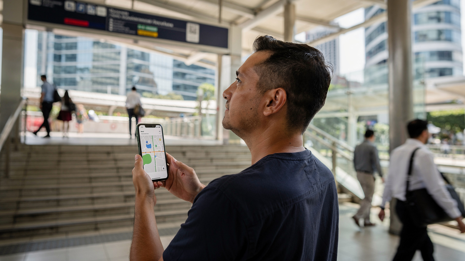 A person holding a phone with a map app open, standing near an MRT station entrance in Singapore, with blurred commuters and iconic city architecture in the background.