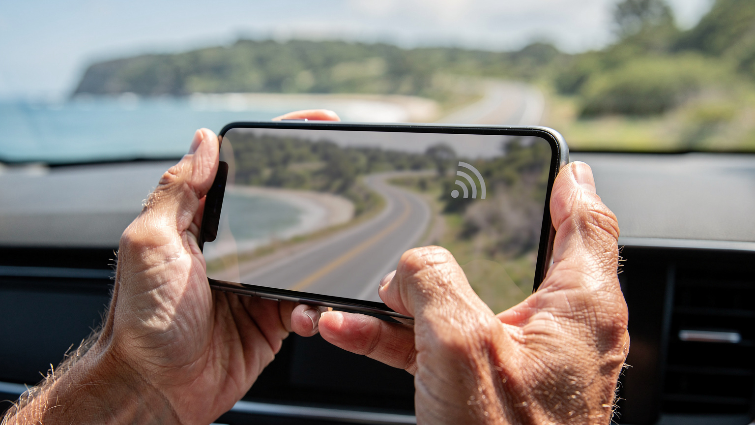 A close-up of a traveller's hands holding a smartphone, with a blurred map interface visible, indicating active navigation on a winding coastal road, suggesting connectivity even in remote areas.