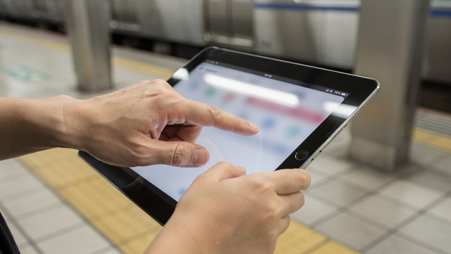 Bir ebeveynin eli, bir çocuğun elindeki iPad'de ebeveyn kontrolü ayarlarını yaparken ekrana dokunuyor, arka planda hafifçe bulanık bir şekilde bir Japon tren istasyonu peronu görünüyor.