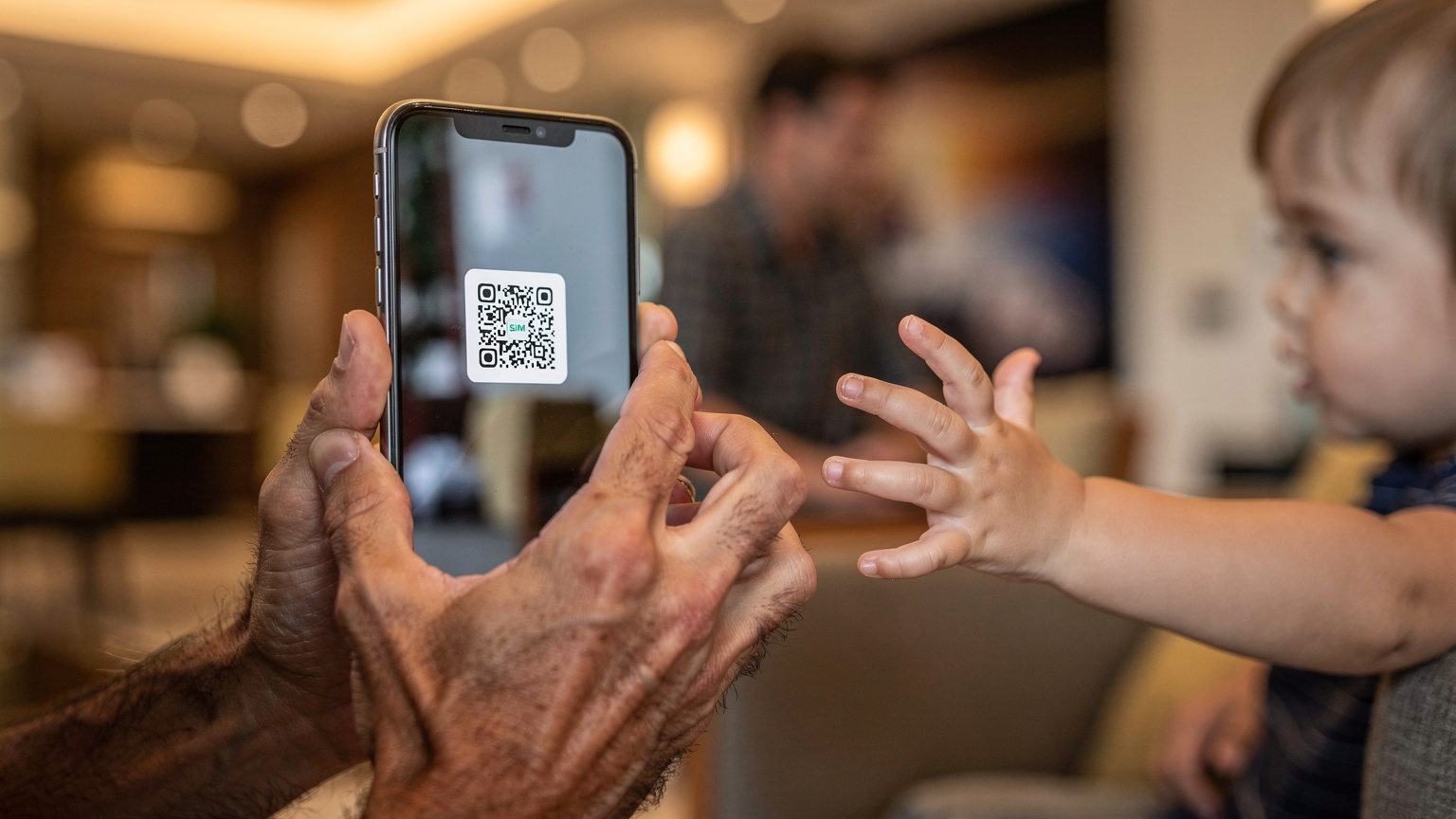 A close-up of a parent's hands, worn from travel, holding a smartphone displaying an eSIM activation QR code, with a child's small hand reaching playfully towards the screen in a hotel lobby.