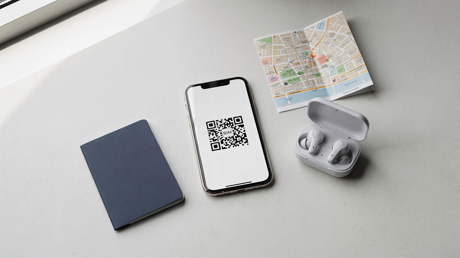 An editorial flat-lay showing a passport, a smartphone with an eSIM QR code on screen, a pair of wireless earbuds, and a small, neatly folded city map, embodying organised business travel essentials.