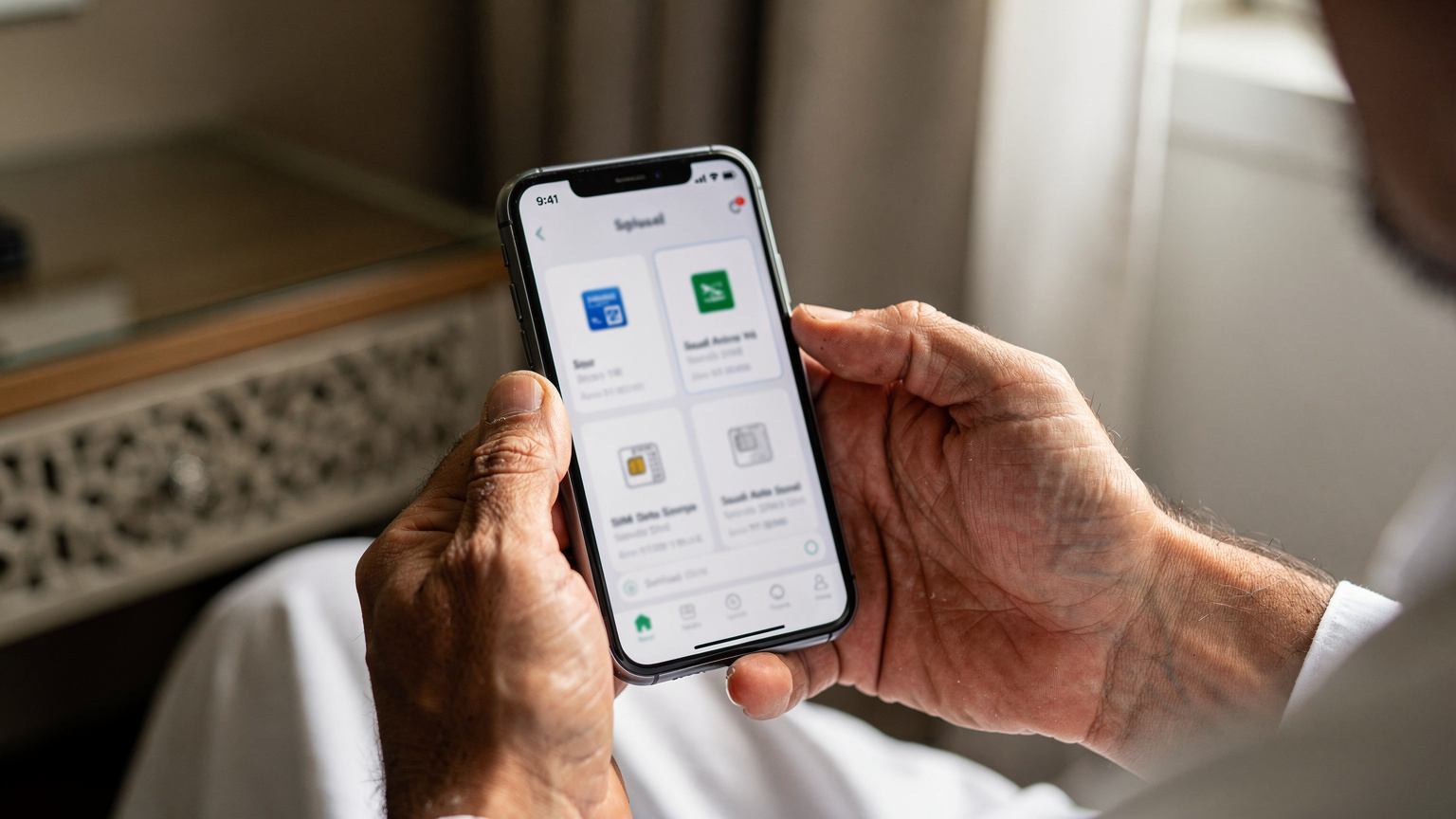 Bir hacı, cep telefonunun ekranında Cellesim uygulamasını açmış, Suudi Arabistan eSIM paketlerini inceliyor. Ekran yakından çekilmiş, ancak metin okunaksız.