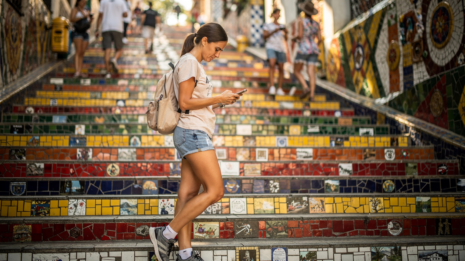 A traveler uses their phone to navigate a bustling street in Rio de Janeiro, with the iconic Escadaria Selarón in the background.