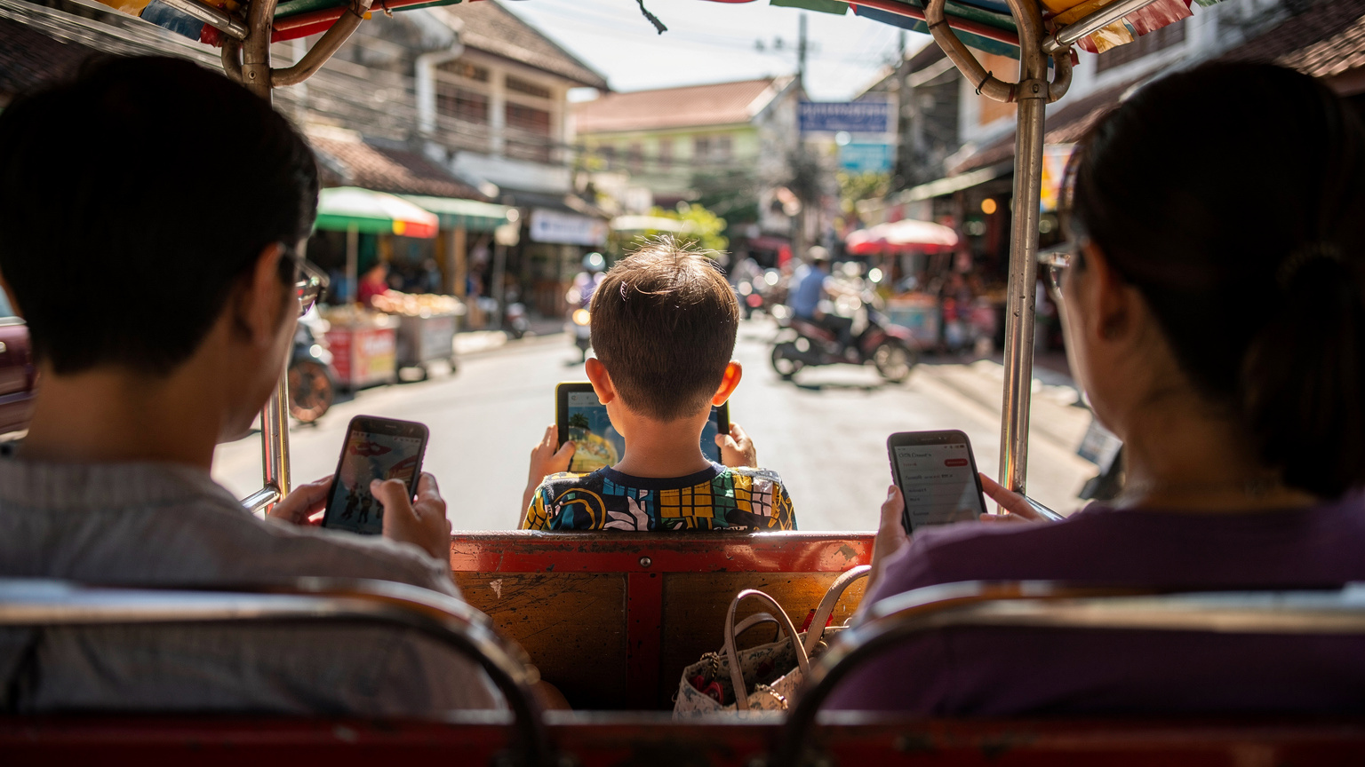 Ein Kind sitzt auf dem Rücksitz eines Tuk-Tuks in Chiang Mai, hält ein Tablet und spielt ein Spiel, während die Eltern im Vordergrund auf ihre Smartphones schauen. Der Hintergrund zeigt eine belebte Straße.