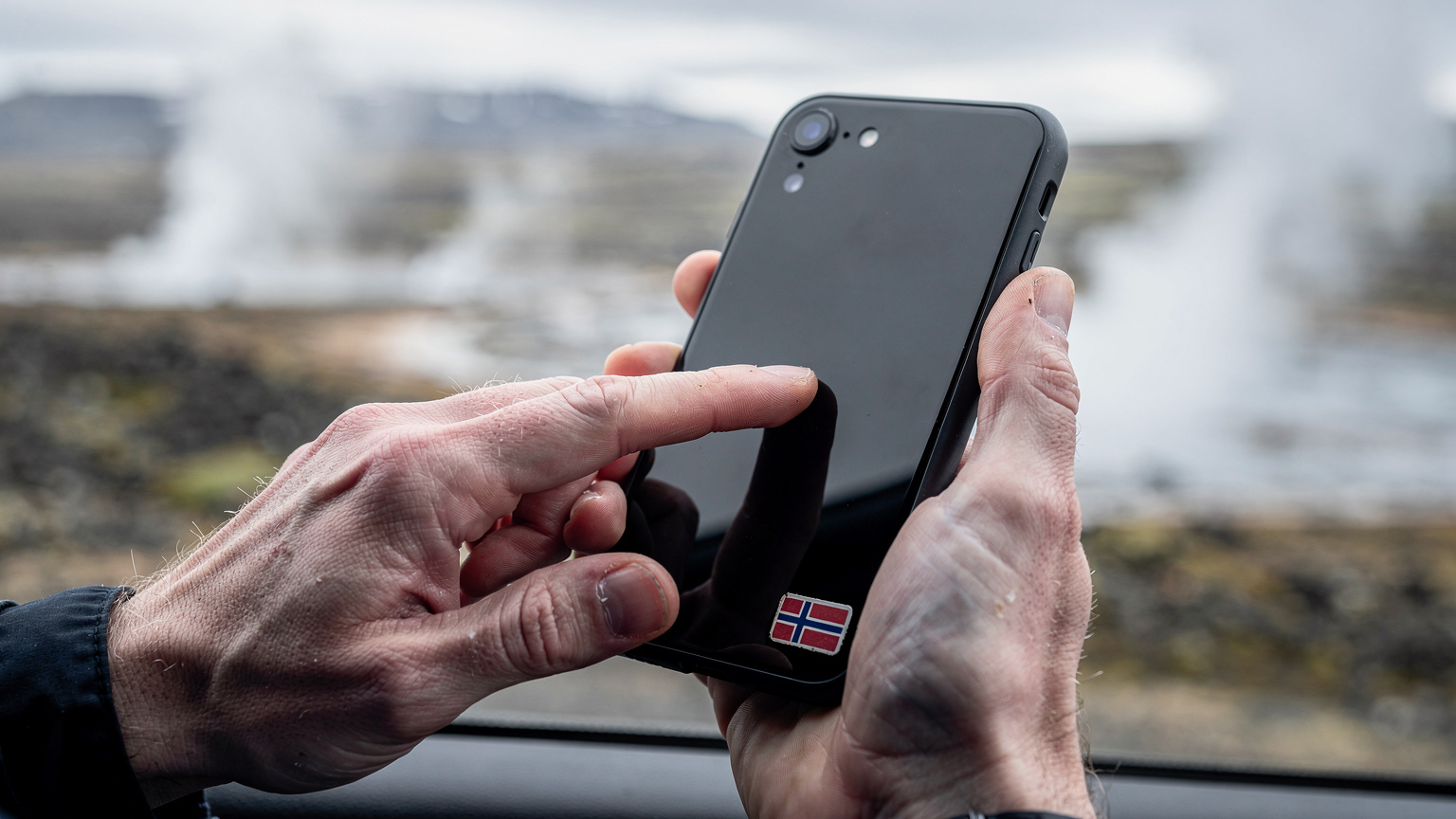 A close-up of a traveler's hands attempting to troubleshoot a smartphone, with a small Icelandic flag sticker visible on the phone case, set against a blurred backdrop of a scenic Icelandic landscape.