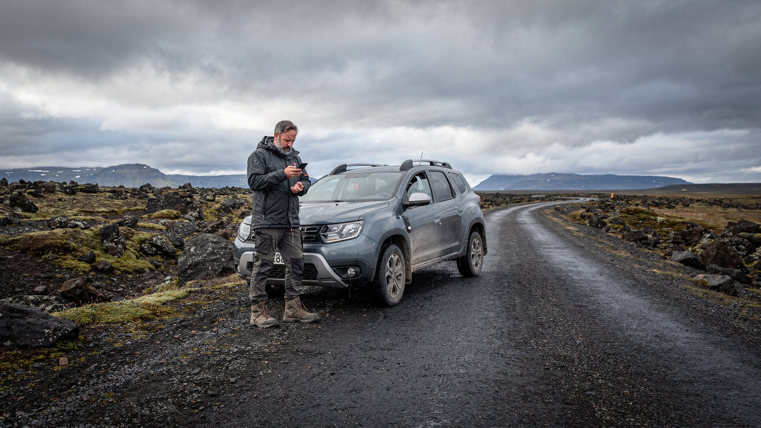 A worried traveler consults their phone, which displays a 'No Signal' message, while standing next to a broken-down car on a desolate Icelandic road, emphasizing the need for reliable connectivity.