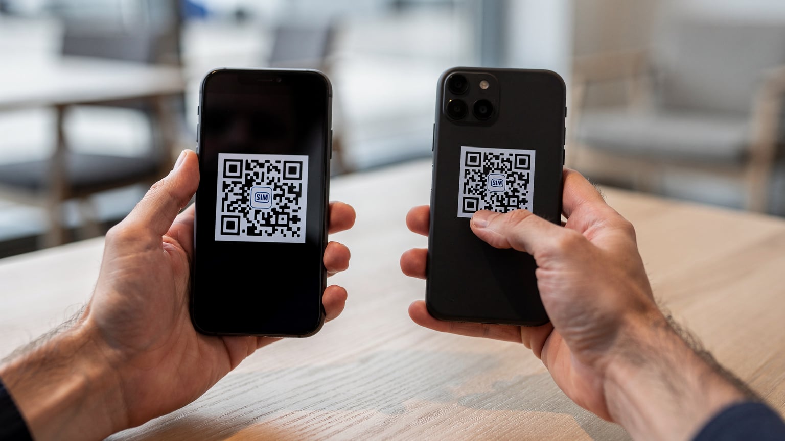 A close-up of a person's hands holding a smartphone, scanning a QR code for eSIM activation, with a blurred background suggesting a travel setting like an airport lounge or hostel common area.