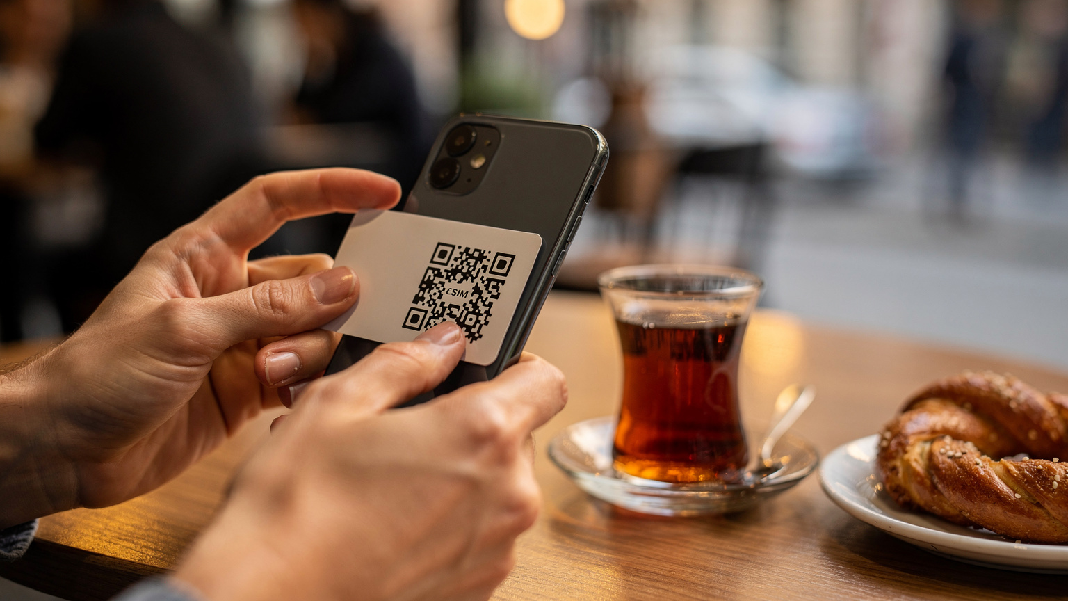Bir seyahat blog yazarının elleri, bir kafede otururken akıllı telefonunda bir eSIM QR kodunu tararken gösteriliyor. Arka planda bulanık bir şekilde bir Türk çay bardağı ve küçük bir tatlı tabağı görülüyor.
