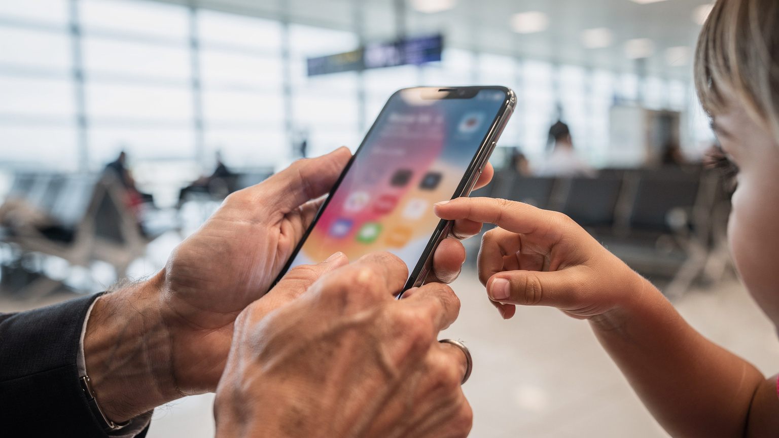 A close-up of a parent's hands expertly navigating an eSIM app on their smartphone, with a child's hand gently touching the screen in curiosity, set against a blurred background of a bustling airport lounge.