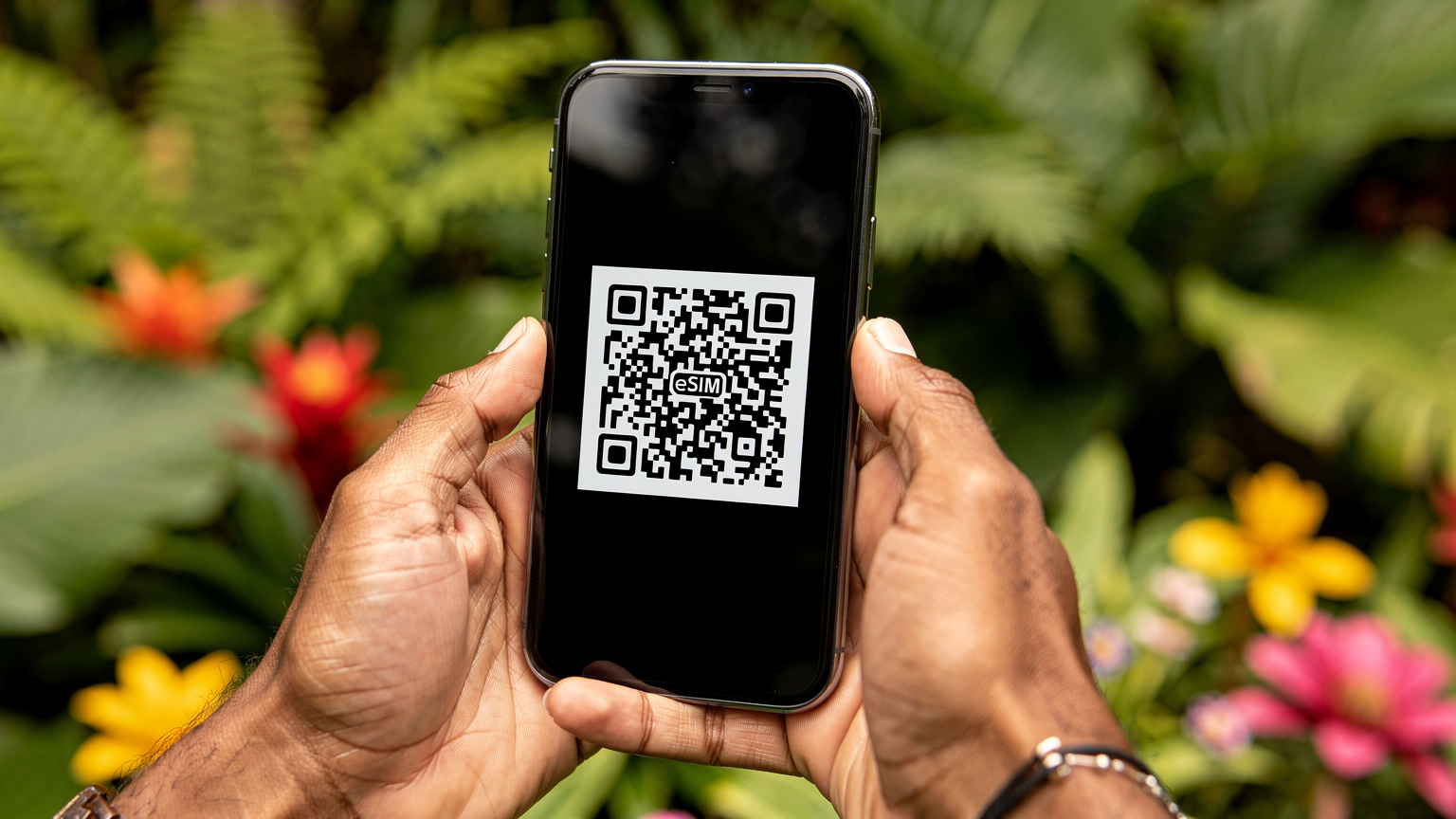 Nahaufnahme von Händen, die ein Smartphone halten, auf dem ein QR-Code für die eSIM-Installation gescannt wird, im Hintergrund verschwimmt ein tropischer Garten.