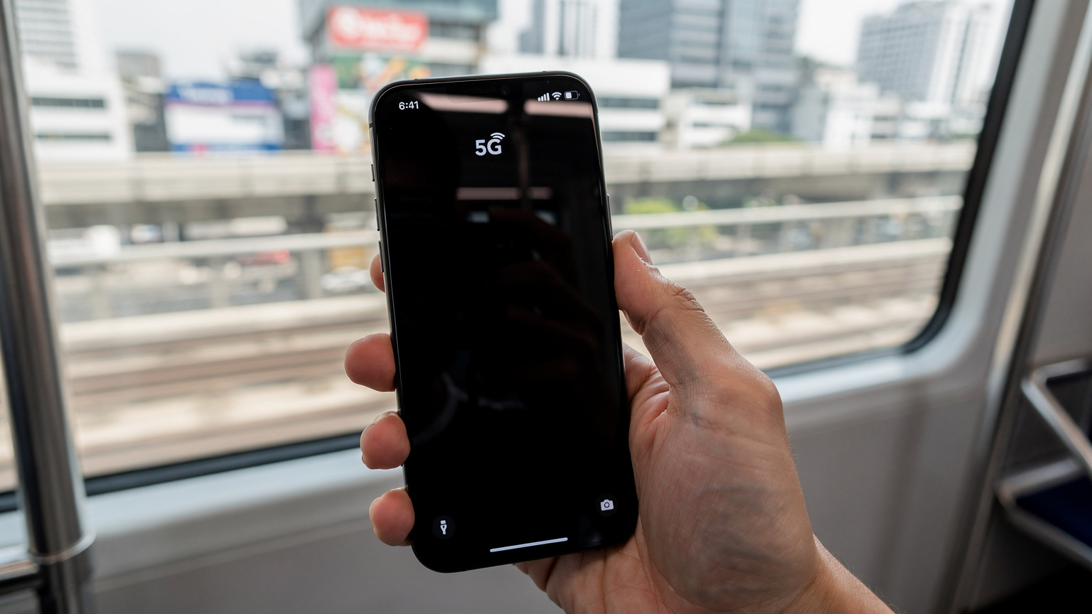 Nahaufnahme einer Hand, die ein Smartphone mit aktivem 5G-Symbol in der oberen Statusleiste hält, während die Person auf einem Skytrain (BTS) in Bangkok fährt.