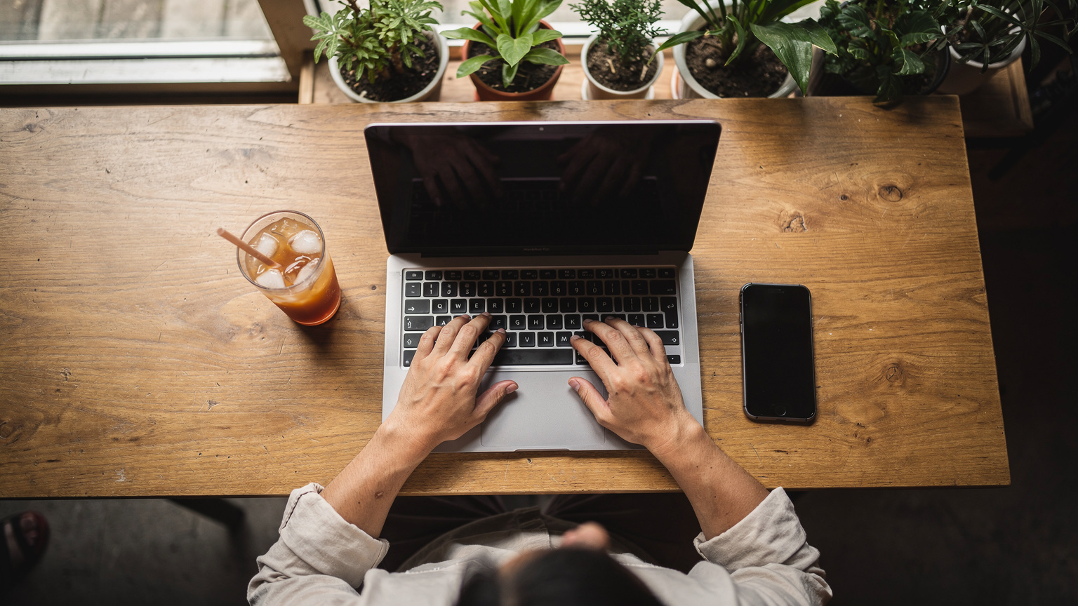 Eine Person sitzt in einem Café in Bangkoks Viertel Ari und arbeitet mit einem Laptop und einem Smartphone, wobei ein traditioneller thailändischer Eiskaffee auf dem Tisch steht.