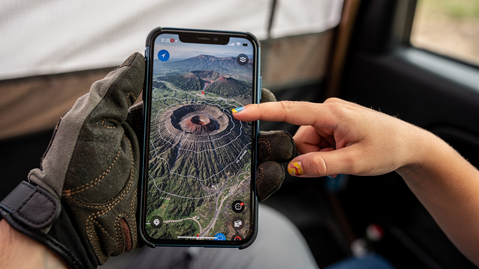 A close-up shot of a parent's hands holding a smartphone, with a child's hand gently touching the screen, looking at a map application displaying a route through a rugged, mountainous landscape.
