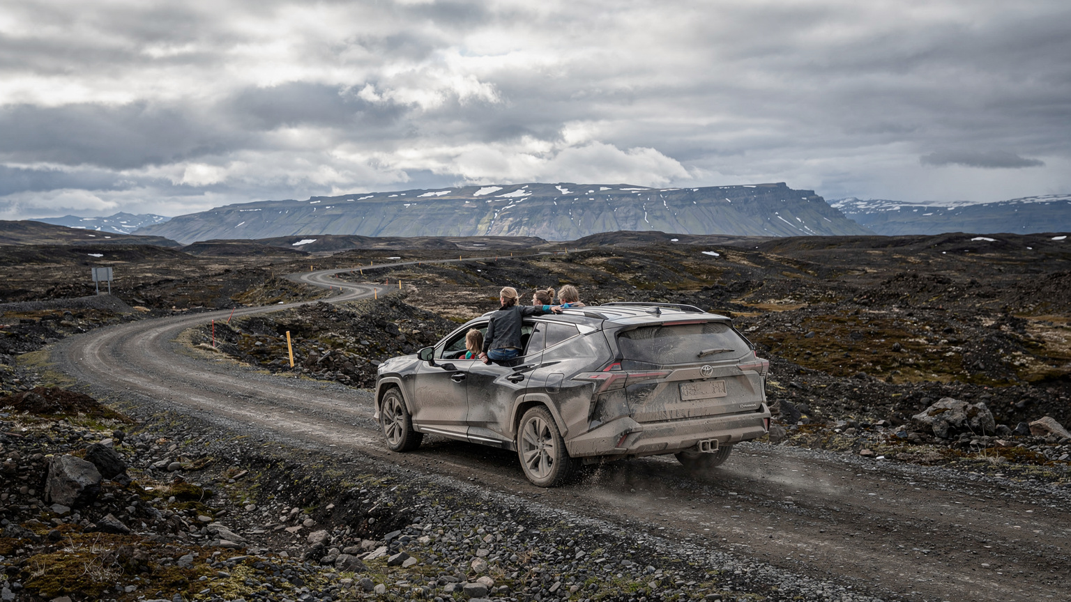 A family of four navigates a rocky, unpaved F-road in the Icelandic Highlands in a 4x4 vehicle, with a vast, empty landscape of volcanic rock and distant mountains under a dramatic sky.