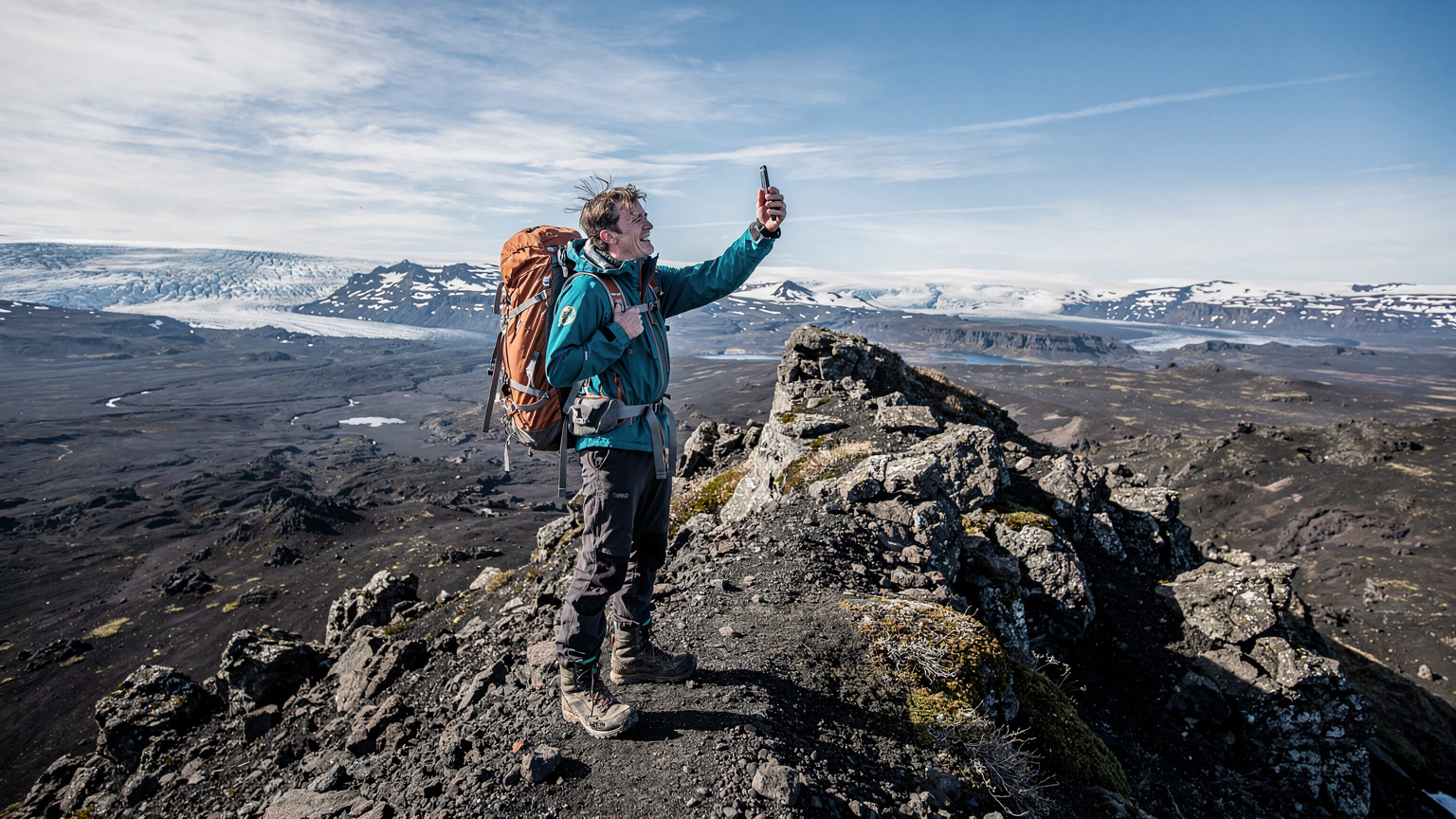 A person in full hiking gear, wearing a backpack, stands on a rugged volcanic ridge in the Icelandic Highlands, holding a smartphone up to try and catch a signal, with a vast, empty landscape stretching behind them.
