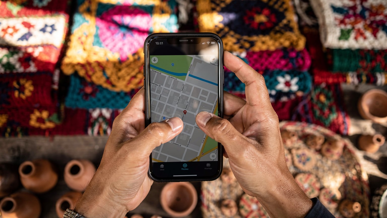 A close-up shot of hands holding a smartphone, displaying a map application, against the backdrop of a vibrant market stall in Oaxaca, Mexico, suggesting travel and local exploration.