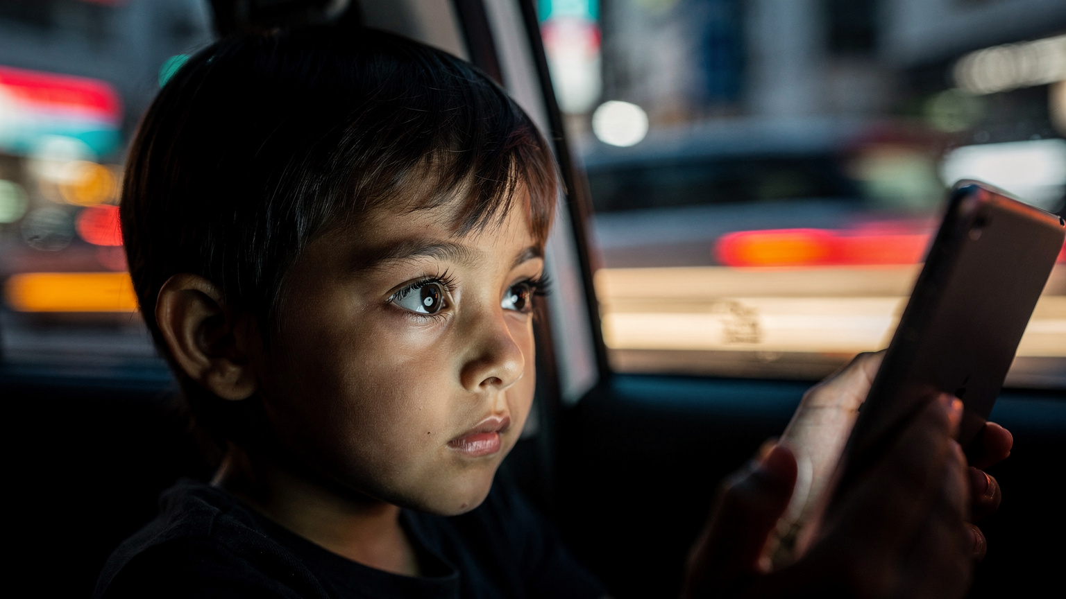 A child looking intently at an iPad, held by an adult hand, while sitting in a car, with blurred city lights outside the window.