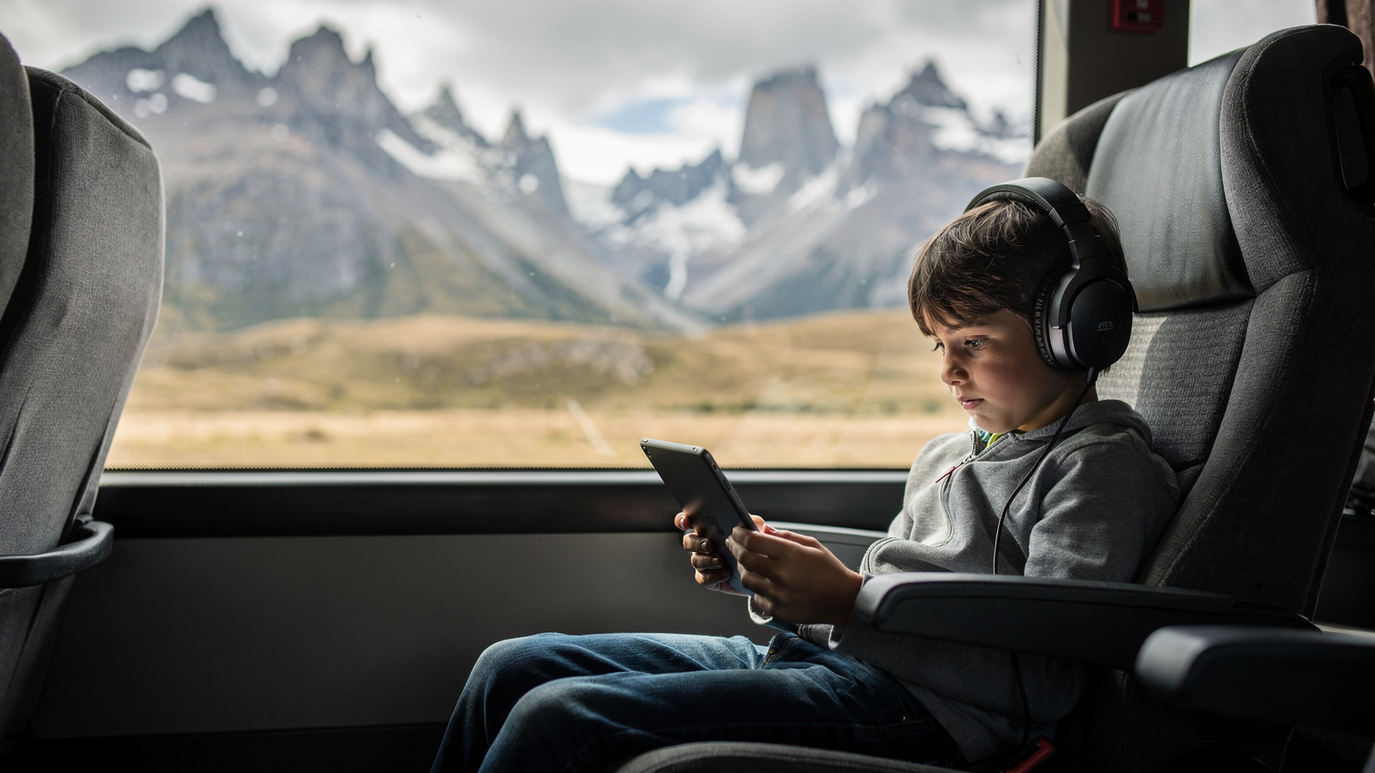 A child, around 9 years old, engrossed in an iPad, with headphones on, sitting comfortably on a long-distance bus in Patagonia. Outside the window, blurred but visible, are sweeping mountain landscapes, illustrating connectivity on the move.