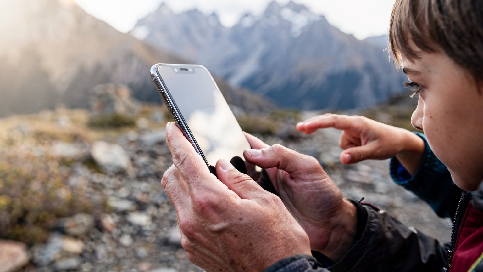 A close-up of a parent's hands, possibly Alex's, demonstrating how to activate an eSIM on a smartphone while standing near a rugged Patagonian mountain trail, with a child's hand briefly reaching into the frame in the background.